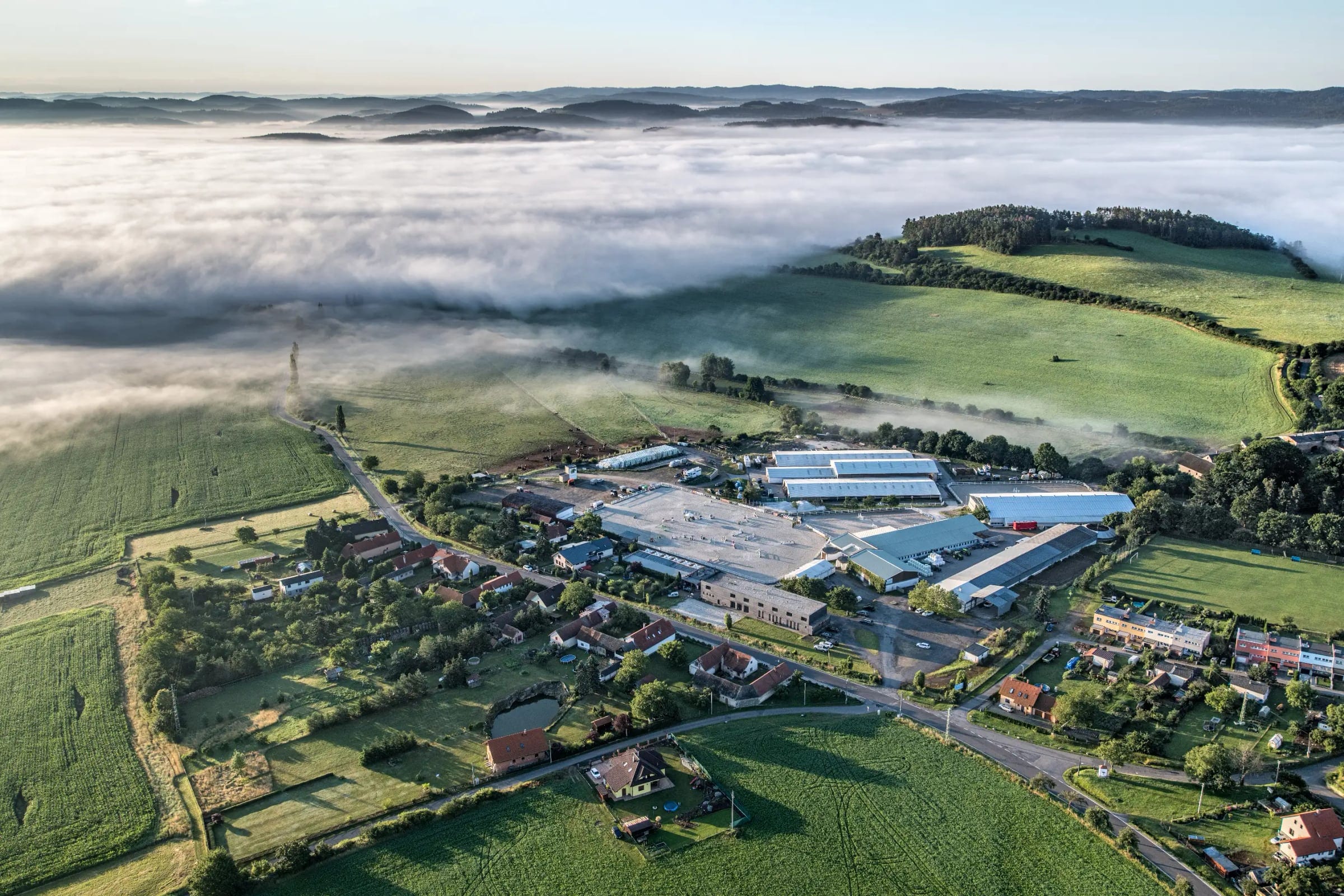 An aerial photo of the equestrian centre at dawn: arenas, stables and halls among green fields, with mist over the hills behind.