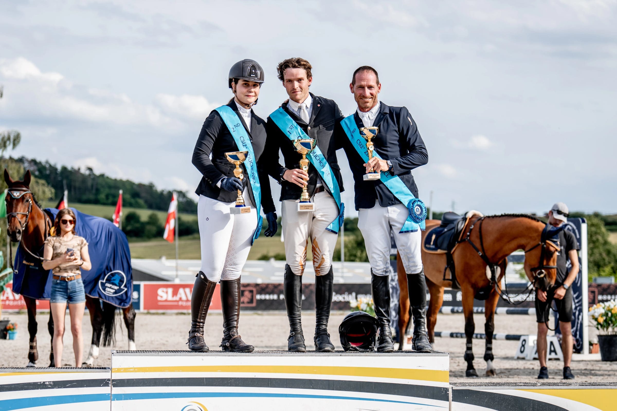 Three riders with trophies and pale blue CSI2* Zduchovice sashes stand on the podium, horses and green hills behind them.