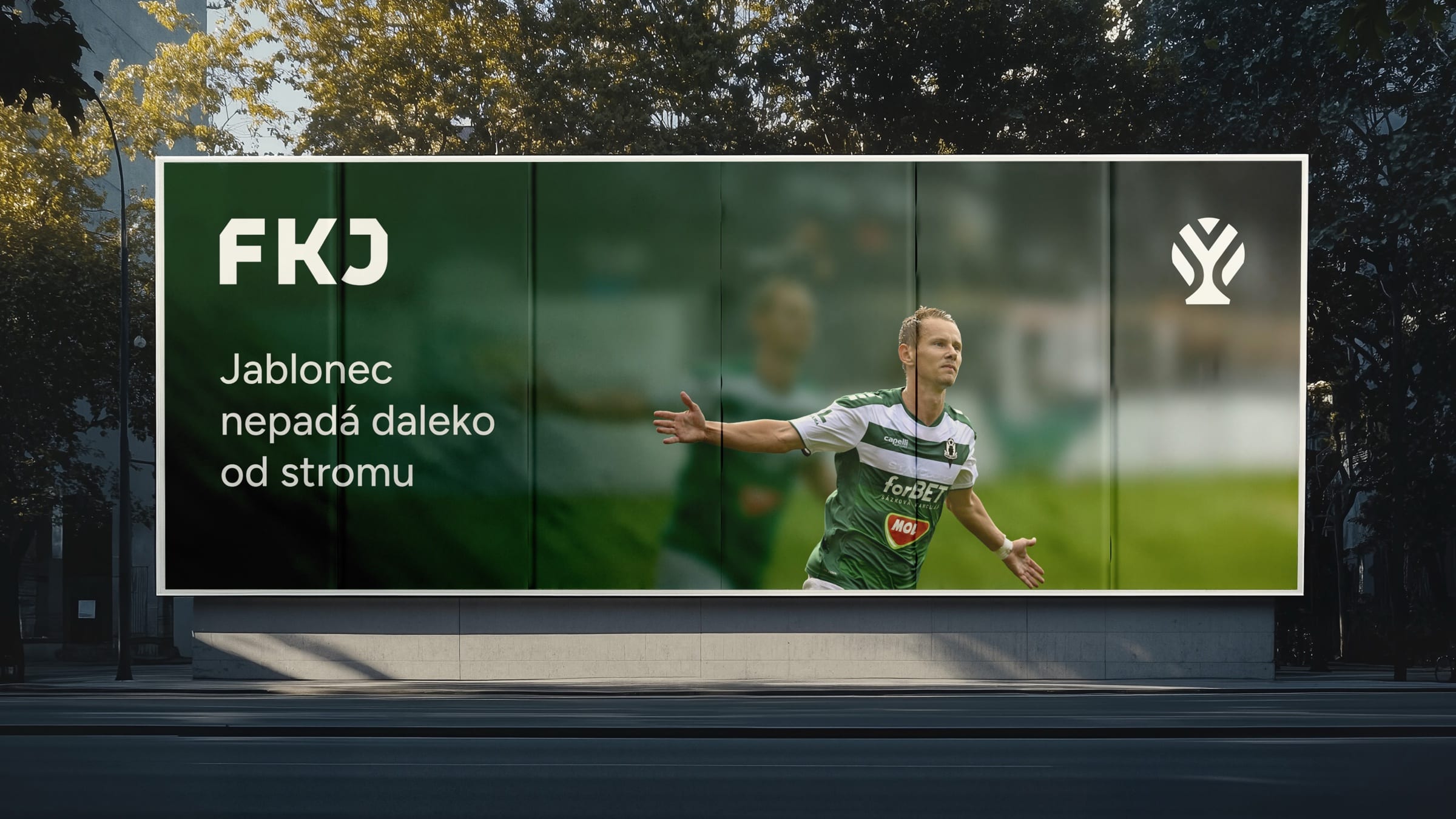 A street billboard with the FKJ wordmark, the apple-tree symbol and a celebrating player, reading “Jablonec nepadá daleko od stromu”.
