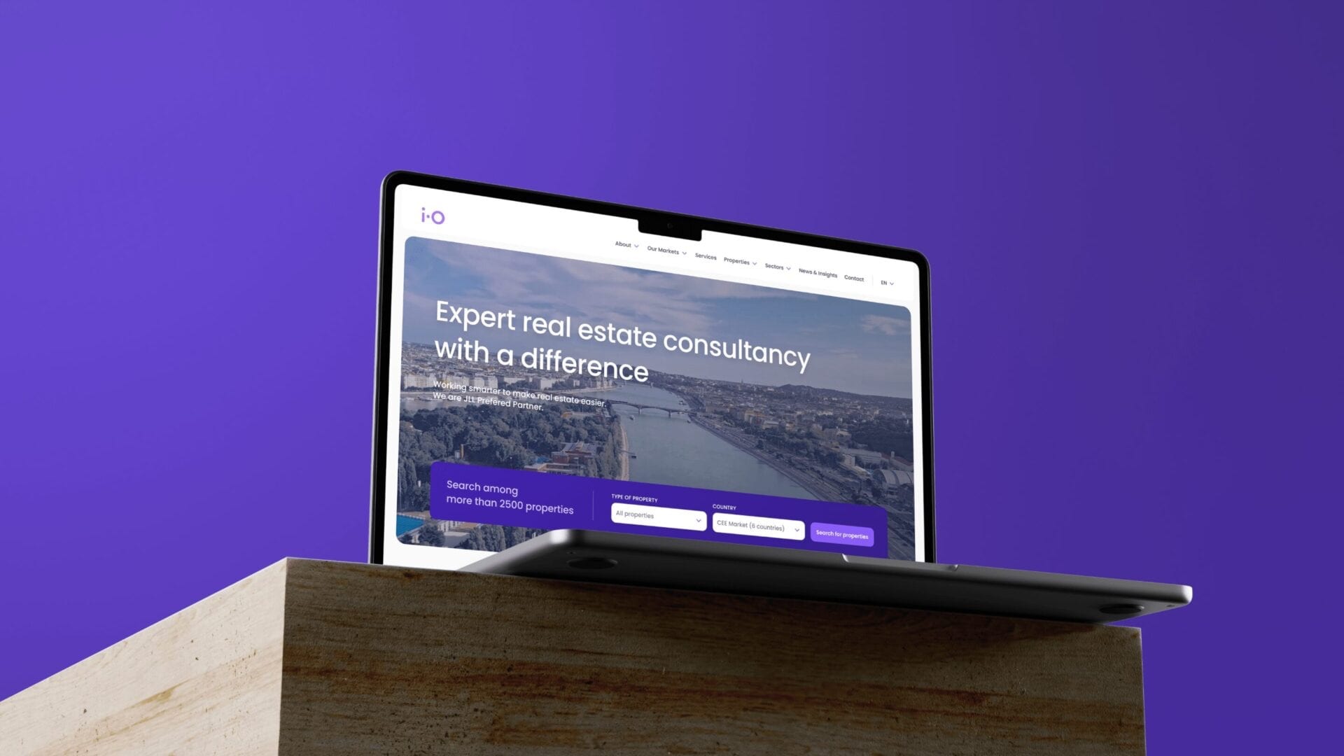 A laptop on a wooden block against violet, showing the iO Partners homepage: “Expert real estate consultancy with a difference” above a property search bar.
