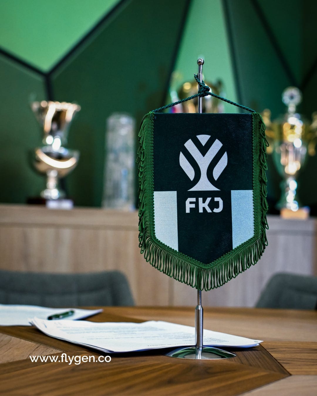 A green club pennant with the FKJ symbol on a table beside trophies.