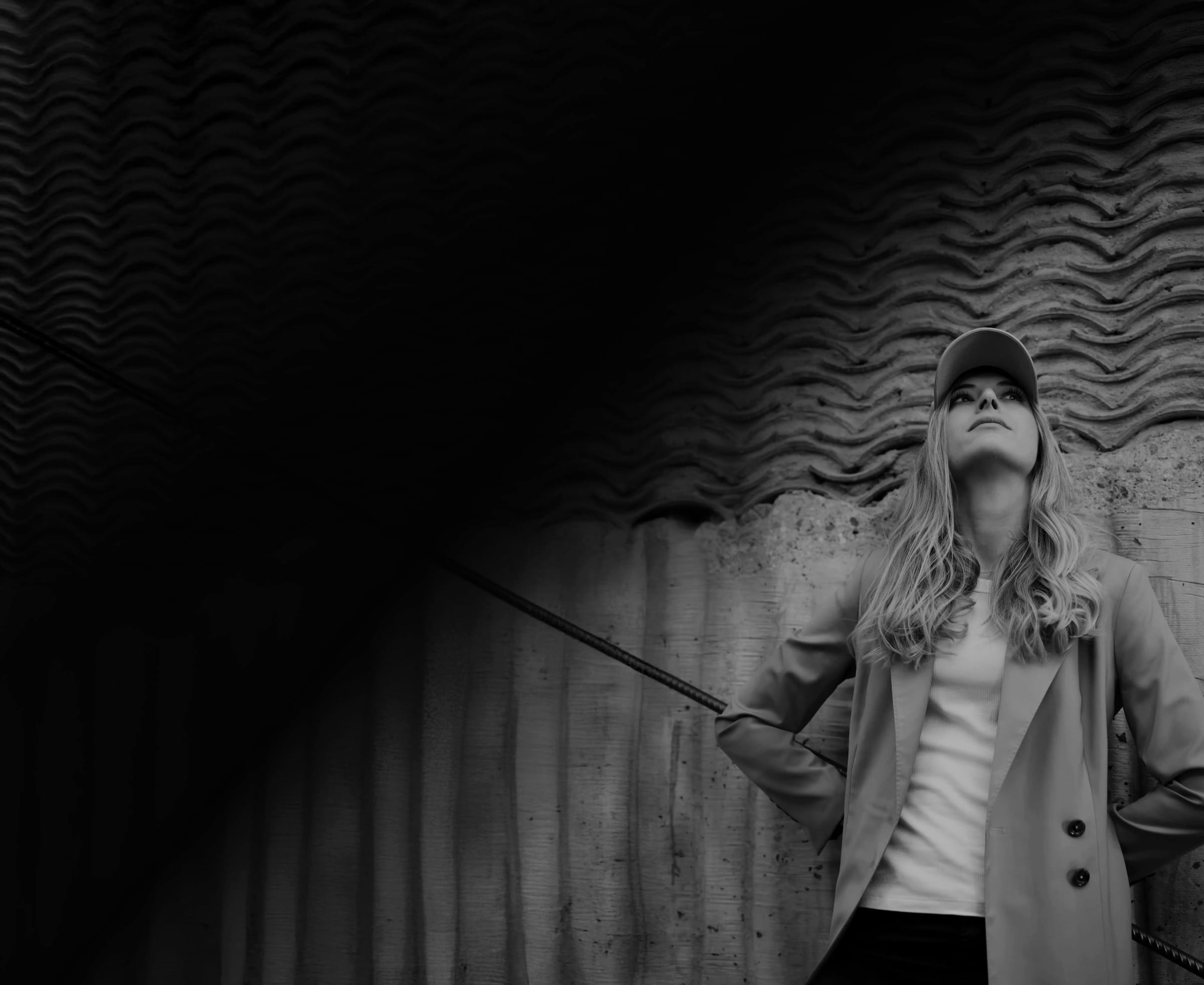 Lucie Krulichová, designer and founder of Flygen, in a cap and long coat against a textured concrete wall, black and white portrait.