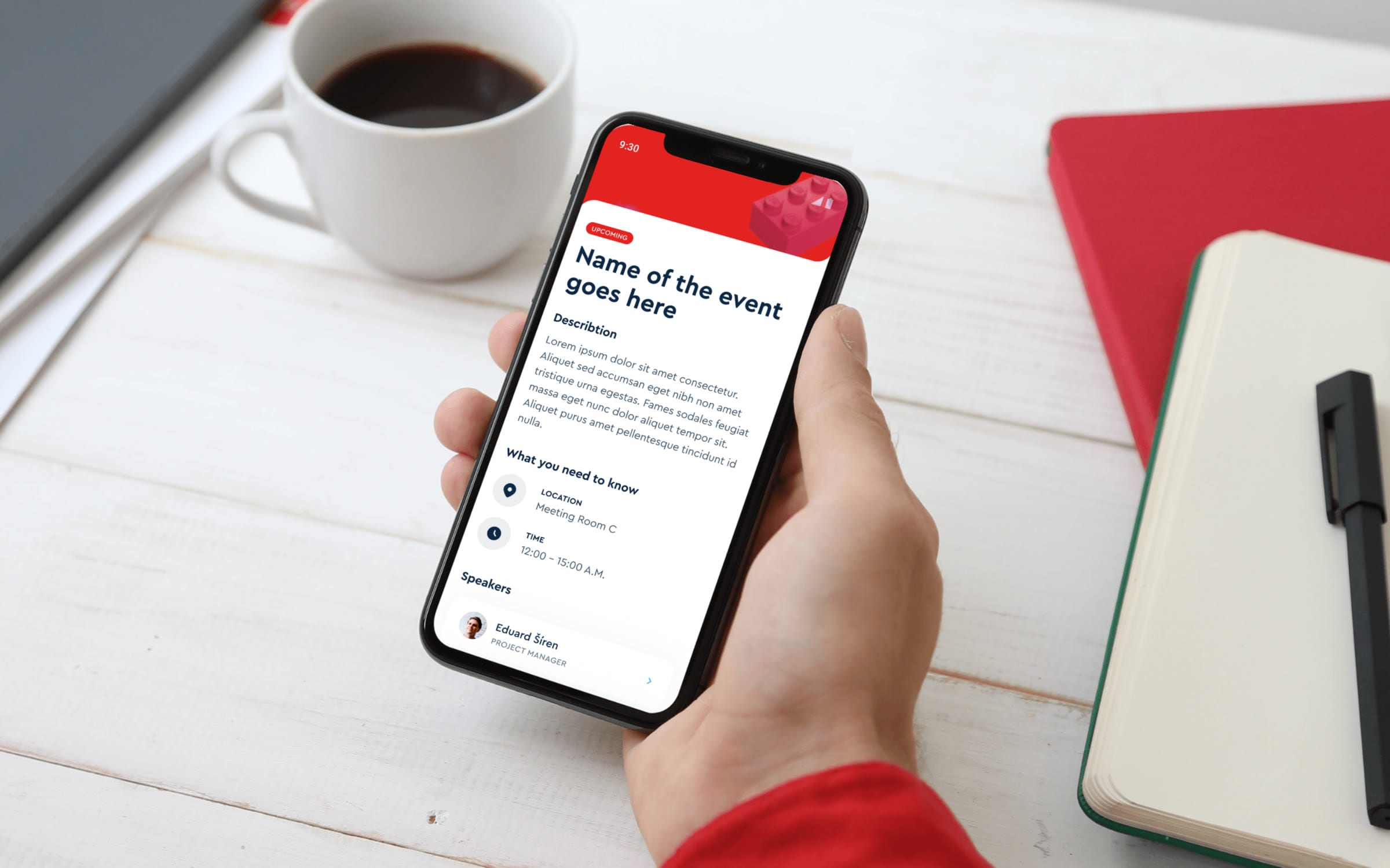 A hand holding a phone with the LEGO event app open on an event page, next to a cup of coffee and a red notebook.